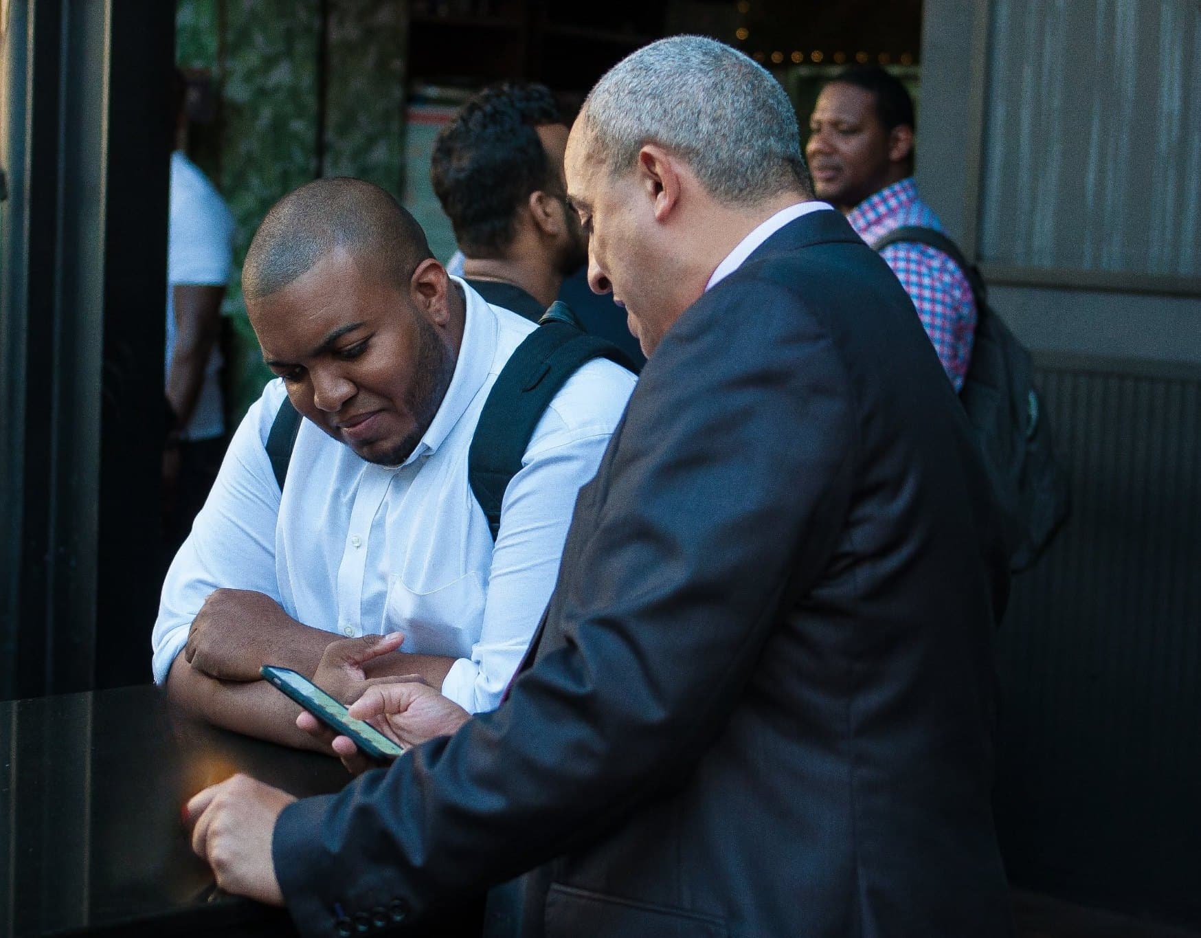 Two men lean over a table looking at a phone together at an evening gathering; one wears a white shirt and a backpack, the other a dark suit.