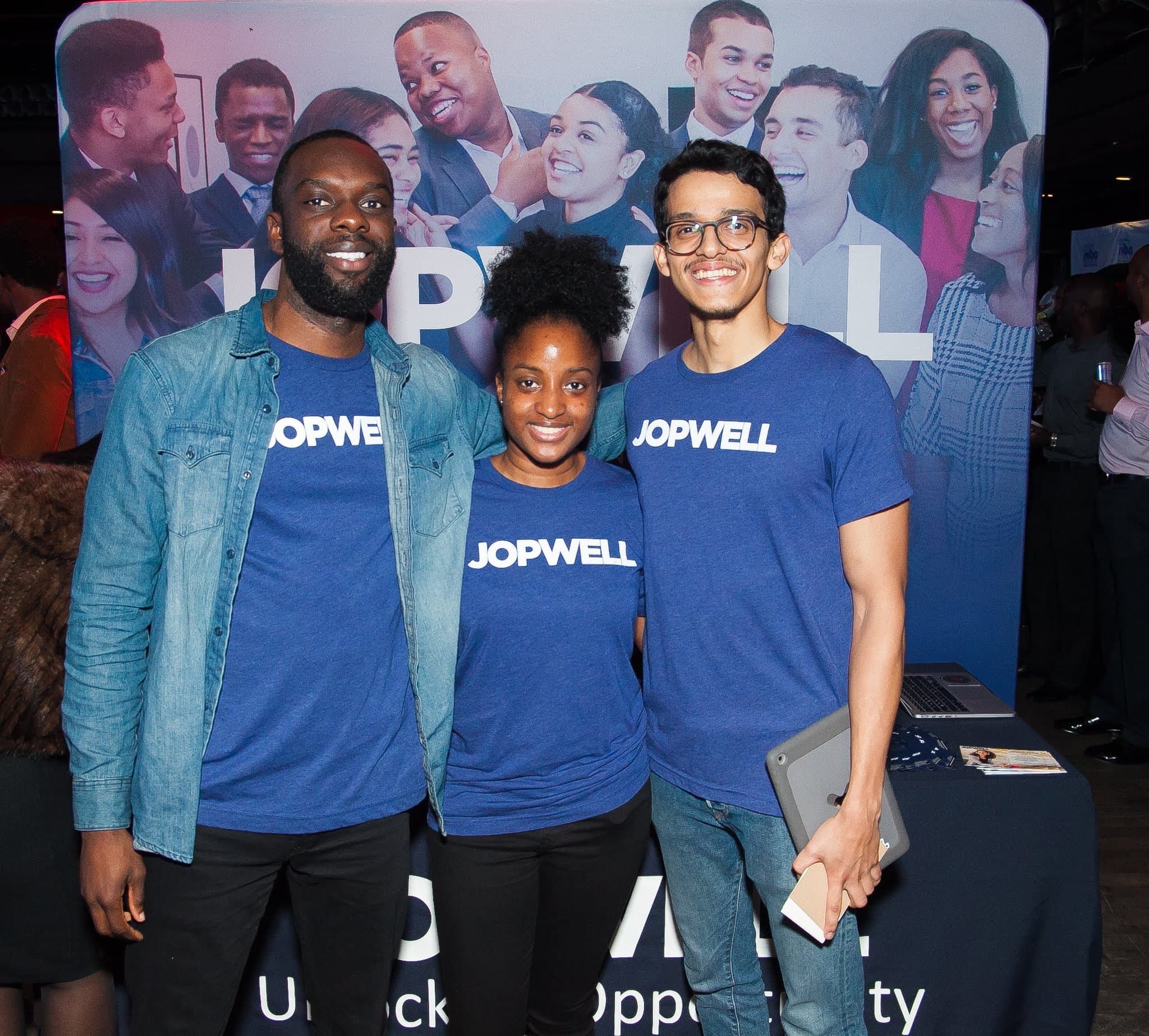 Three smiling people wearing blue Jopwell T-shirts pose together in front of a Jopwell banner.