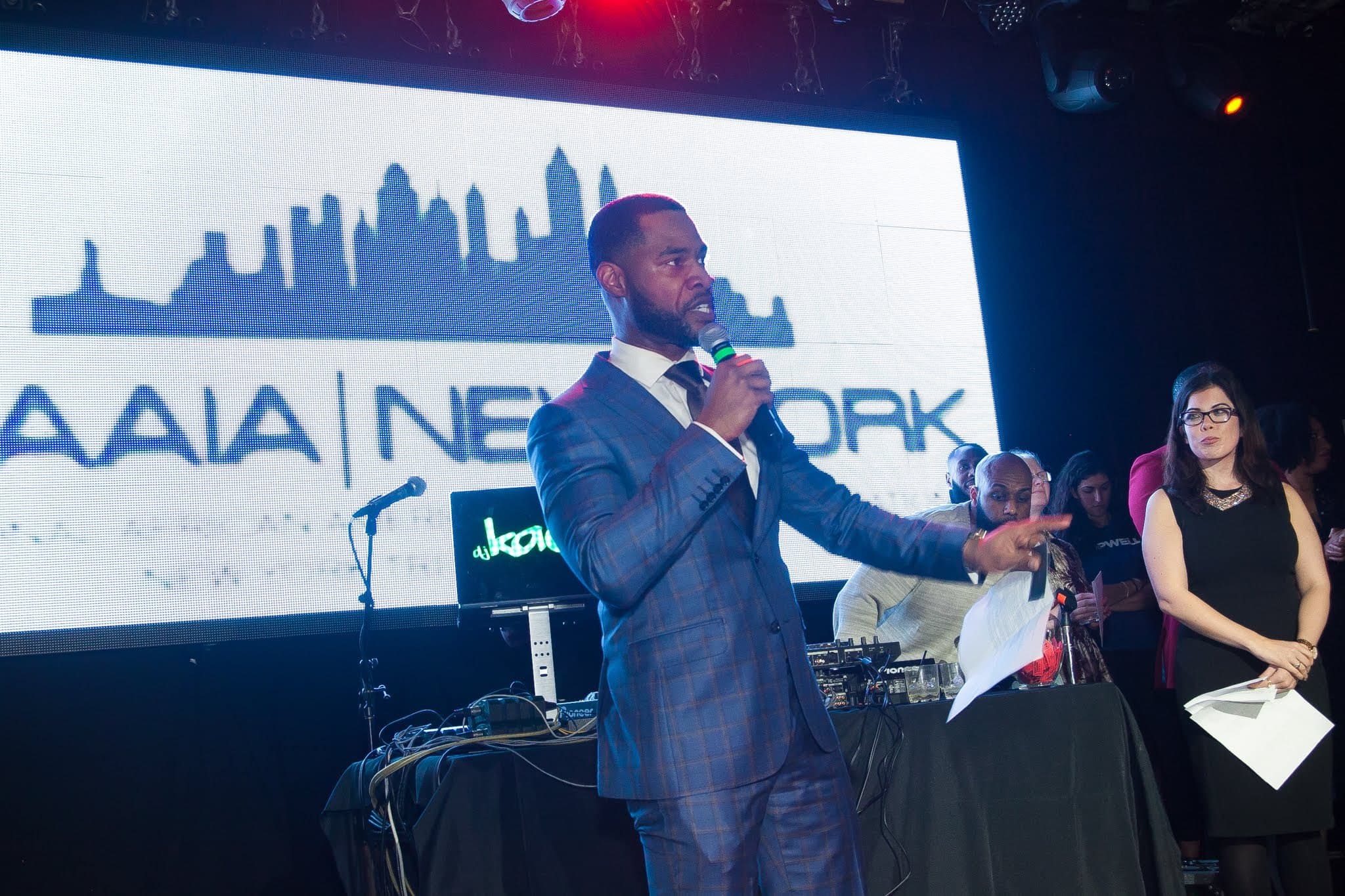A man in a blue suit speaks into a microphone on stage in front of a large screen while a woman holding papers looks on.