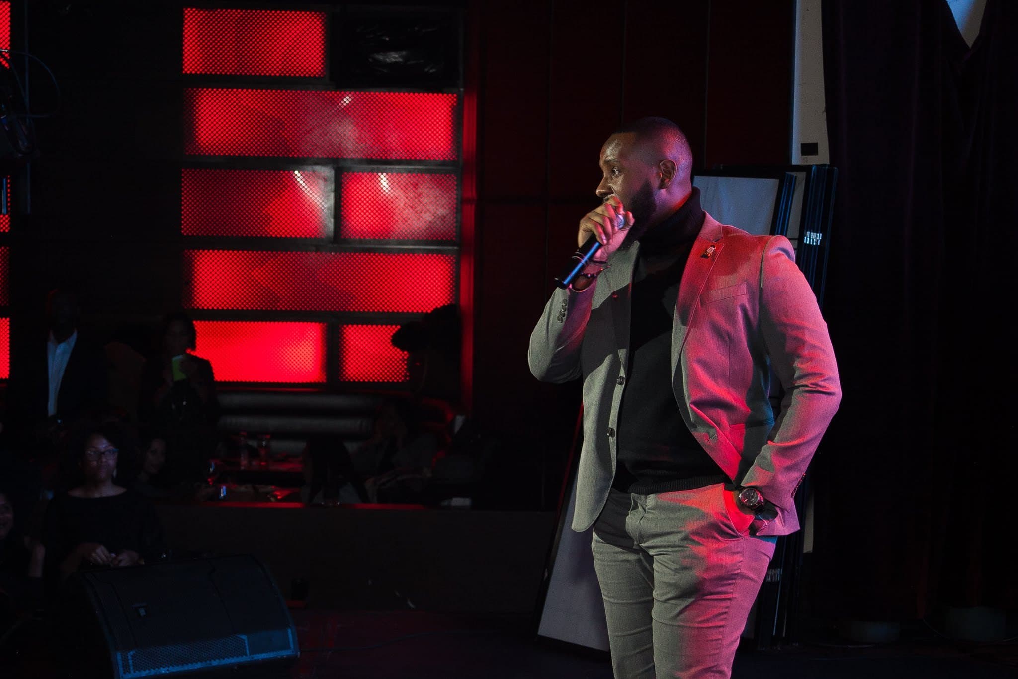 A man in a grey coat speaks into a microphone on a stage lit in red.